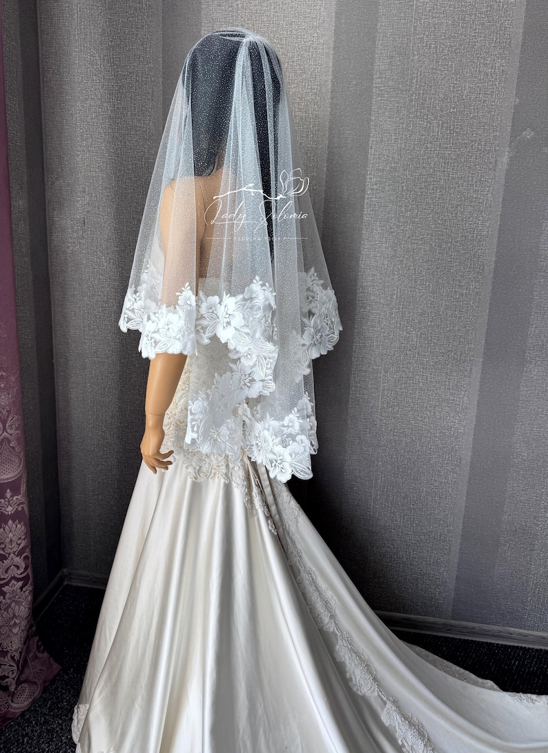 May include: A white wedding veil featuring a shimmering tulle fabric and a wide border of intricate floral lace appliqu&eacute;. The veil is draped over a mannequin wearing a floor-length satin gown with a train. The text Lady Alonia is visible.