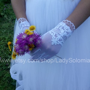 Puede incluir: Guantes blancos transparentes hasta la mu&ntilde;eca con intrincados bordados de encaje floral y borde festoneado. Los guantes se llevan con un vestido de tul blanco y sostienen un peque&ntilde;o ramo de flores silvestres moradas y amarillas. Texto: www.etsy.com/shop/LadySolomia.