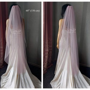 May include: A long, sheer, dusty rose-colored wedding veil made of soft tulle fabric. The veil measures 60 inches in length and is draped over a mannequin wearing a white satin gown with lace detailing. The Lady Sabina logo is visible on the veil.