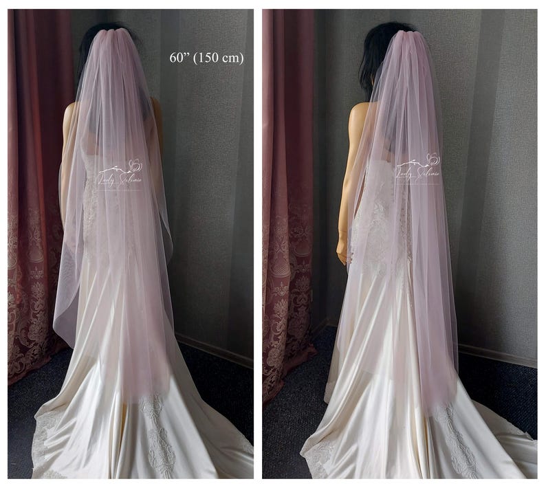 May include: A long, sheer, dusty rose-colored wedding veil made of soft tulle fabric. The veil measures 60 inches in length and is draped over a mannequin wearing a white satin gown with lace detailing. The Lady Sabina logo is visible on the veil.