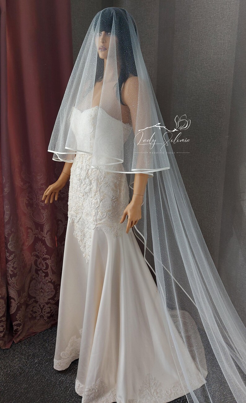 May include: A white bridal veil with a delicate sparkle and a satin trim. The veil is layered and has a long, flowing train.
