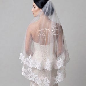 May include: A two-tier white bridal veil featuring a delicate glitter tulle fabric and a scalloped floral lace trim along the edges. The veil is draped over the head and shoulders, showcasing a sheer, shimmering texture and intricate lace detailing.