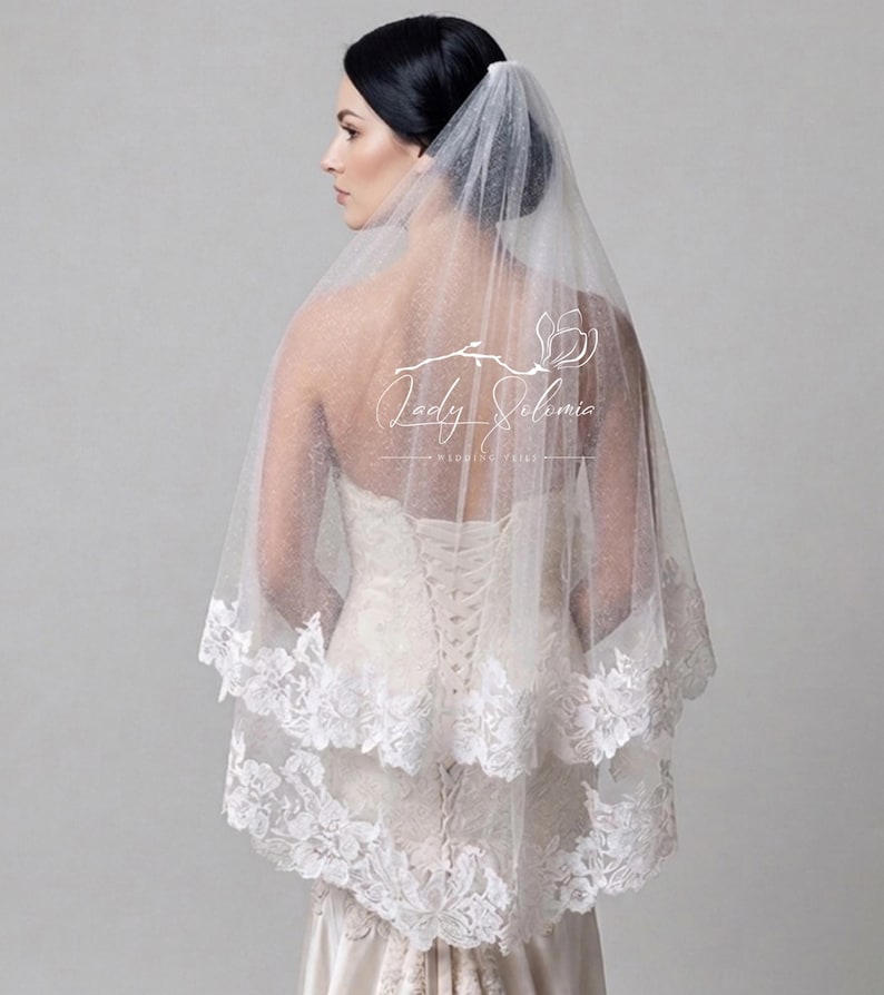 May include: A two-tier white bridal veil featuring a delicate glitter tulle fabric and a scalloped floral lace trim along the edges. The veil is draped over the head and shoulders, showcasing a sheer, shimmering texture and intricate lace detailing.