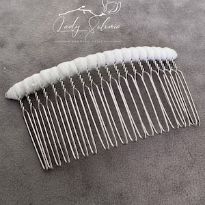 Puede incluir: Una peineta de metal plateado con el borde superior curvo envuelto en tela blanca. La peineta tiene veintidós dientes largos, finos y redondeados. Sobre la peineta, el texto Lady Solomia Wedding Veils está impreso en color blanco.