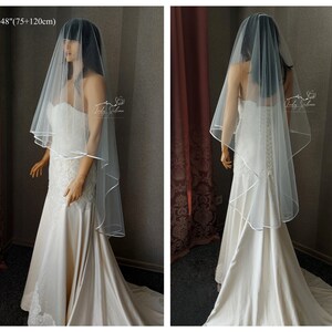 May include: A white bridal veil with a satin edge. The veil is 30 inches long and has a 48 inch drop. The veil is shown on a mannequin wearing a white wedding dress.