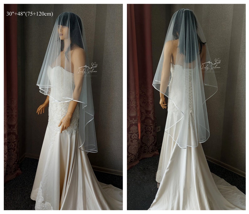 May include: A white bridal veil with a satin edge. The veil is 30 inches long and has a 48 inch drop. The veil is shown on a mannequin wearing a white wedding dress.