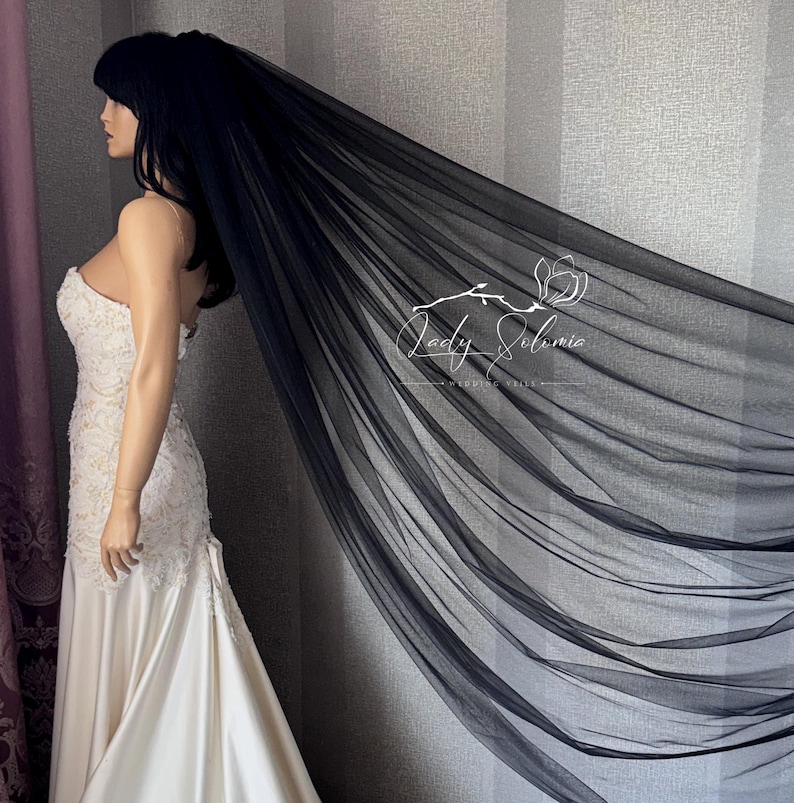 May include: A long, sheer black wedding veil made of fine tulle fabric, draped elegantly to show its length and transparency. The veil features the white logo text Lady Solomia printed on the fabric. It is paired with a white lace wedding dress.