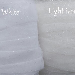 Puede incluir: Dos rollos de tela de tul transparente con purpurina mostrados uno al lado del otro. El rollo izquierdo est&aacute; etiquetado como White y el derecho como Light ivory. Ambas telas presentan una textura de malla fina y transl&uacute;cida con destellos.