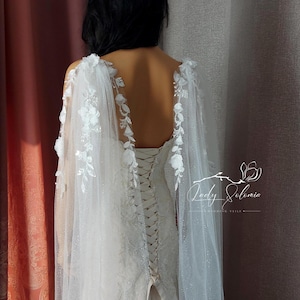 May include: A white wedding dress with a lace-up back and a sheer, floral-adorned cape. The cape features delicate white flowers and extends over the shoulders. The dress is detailed with intricate lace patterns. The image includes the text "Lady Silvia Wedding Veils."