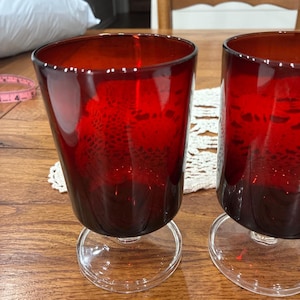 May include: Two deep red glass goblets with a smooth, rounded bowl shape and a clear glass stem and circular base. A pink measuring tape is visible in the background, showing a measurement of approximately 4 inches on the visible segment.