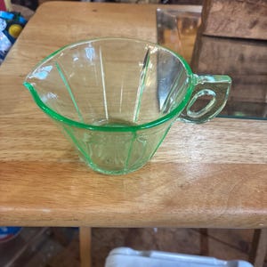 May include: A vintage green glass creamer with a handle and a spout. The creamer is made of a light green glass and has a geometric design. It is sitting on a wooden table.