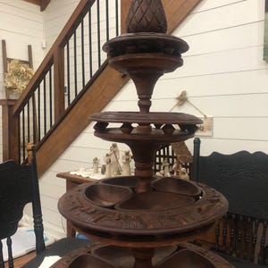 Pode incluir: Um suporte de servir de madeira castanha escura em camadas com um pinheiro no topo. O suporte tem várias bandejas circulares com detalhes esculpidos e seções divididas, ideal para exibir alimentos ou itens decorativos. Uma peça única de decoração.