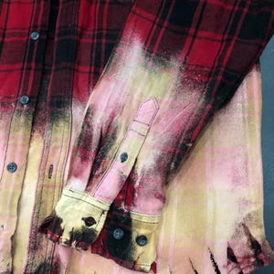May include: Red and black plaid button-down shirt with a bleached, distressed effect. The sleeves and lower portion of the shirt have a faded, pinkish-yellow hue. Features a classic collar, buttoned cuffs, and a chest pocket.