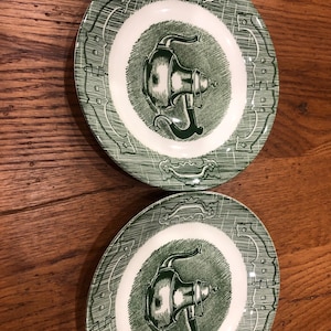 May include: Two green and white decorative plates. Each plate features a central design of a vintage-style machine, surrounded by a white border and a green patterned rim. The plates are displayed on a wooden surface.