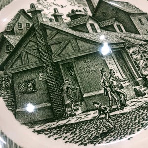 Może przedstawiać: Biały ceramiczny talerz z zielonym wzorem transferowym. Wzór przedstawia szczegółową scenę uliczną z budynkami, postaciami i tekstem "The Old Curiosity Shop". Talerz ma falisty brzeg.
