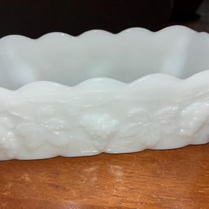May include: A white, rectangular dish with a scalloped rim. The sides are decorated with a grape and leaf pattern. The dish is made of a glossy, opaque material, likely glass or ceramic.