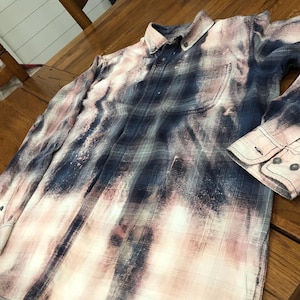 May include: A long-sleeved button-down shirt with a plaid pattern in shades of navy blue, pink, and white. The shirt has a bleached or distressed effect, with a pocket on the chest and buttoned cuffs. The shirt is laying on a wooden surface.