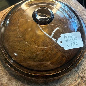 Puede incluir: Tapa de vidrio Pyrex V15C de color ámbar con un pomo negro. La tapa redonda y transparente tiene un ligero tinte ámbar. Una etiqueta blanca adjunta a la tapa dice "Pyrex AMBER V15C".