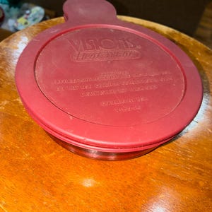 Könnte beinhalten: Roter Deckel für Visions Heat n' Eat Behälter. Der Deckel hat einen Griff und den Text "VISIONS Heat n' Eat". Der Deckel ist rund und aus strapazierfähigem Material gefertigt. Für Kühlschrank, Gefrierschrank und Mikrowelle geeignet.