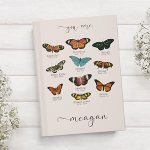 May include: A pink journal with the words "you are" and "meagan" written in cursive. The journal features a collage of colourful butterflies with bible verses printed below each butterfly.