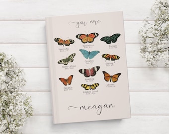 May include: A pink journal with the words "you are" and "meagan" written in cursive. The journal features a collage of colorful butterflies with bible verses printed below each butterfly.
