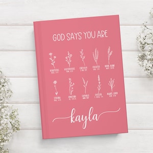 May include: A pink journal with the text "GOD SAYS YOU ARE" in white lettering. The journal has a list of positive affirmations with bible verses in white lettering. The name "kayla" is written in cursive at the bottom of the journal.