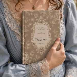 May include: A hardback book with a taupe floral pattern cover featuring an ornate cream-coloured frame. The text reads: His Word Written in My Heart, Victoria, and Psalm 119:11. The book is held against a light blue garment with lace trim.