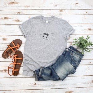 May include: Gray t-shirt with the text "gigi est. 2022" in black script font. The shirt is laid flat on a white wood surface with a pair of brown sandals and blue jeans.