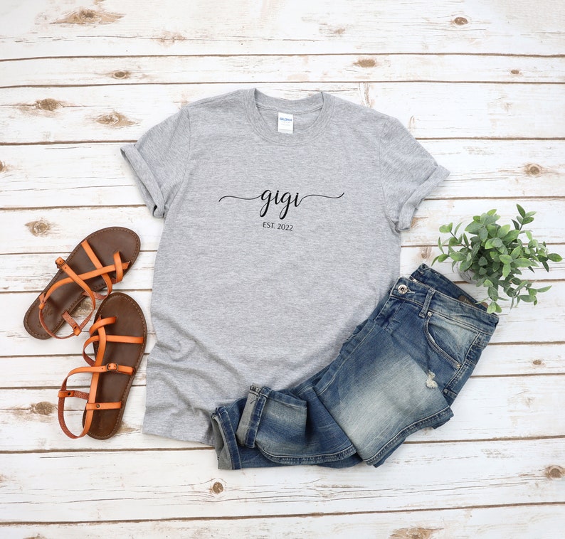 May include: Gray t-shirt with the text "gigi est. 2022" in black script font. The shirt is laid flat on a white wood surface with a pair of brown sandals and blue jeans.