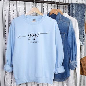 May include: Light blue sweatshirt with black text that reads "gigi est. 2022" hanging on a rack.