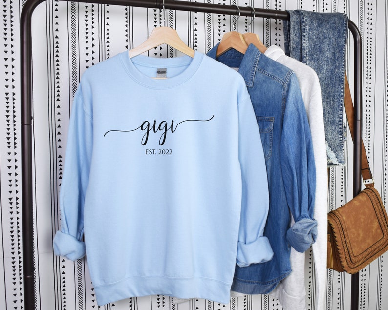 May include: Light blue sweatshirt with black text that reads "gigi est. 2022" hanging on a rack.