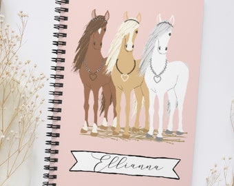 May include: A pink spiral bound notebook with a personalized name "Ellianna" on a banner. The cover features a whimsical illustration of three horses with different colored manes and tails, wearing jeweled necklaces.