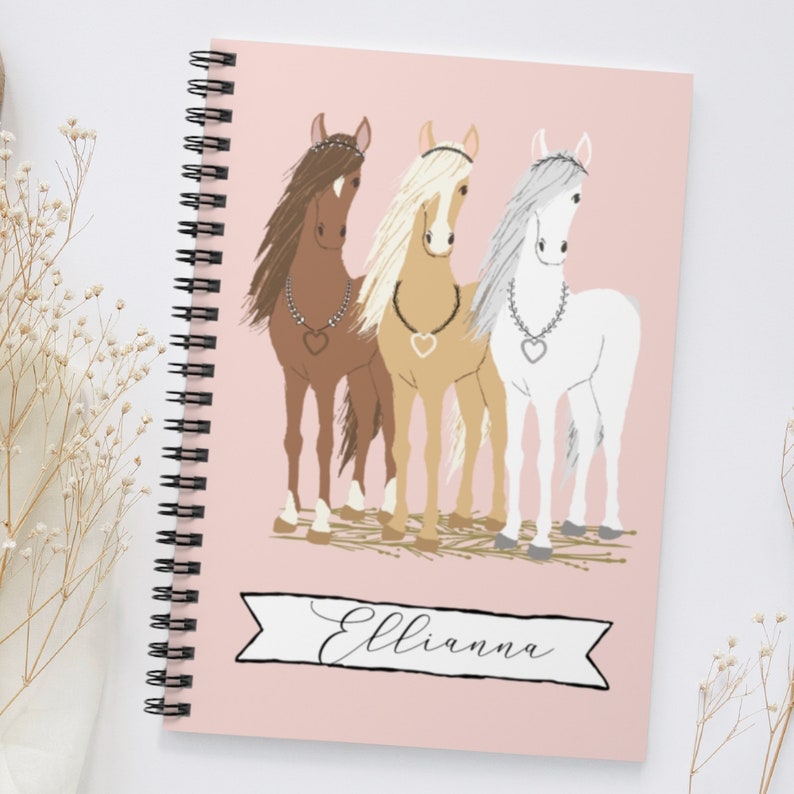May include: A pink spiral bound notebook with a personalized name "Ellianna" printed on a banner. The cover features a whimsical illustration of three horses with different colored manes and tails, wearing jeweled necklaces.
