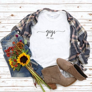 May include: White t-shirt with the text "gigi est. 2022" in black script font. The shirt is laid out on a white wood background with a pair of blue jeans, a plaid shirt, a bouquet of sunflowers and other flowers, and a pair of brown ankle boots.