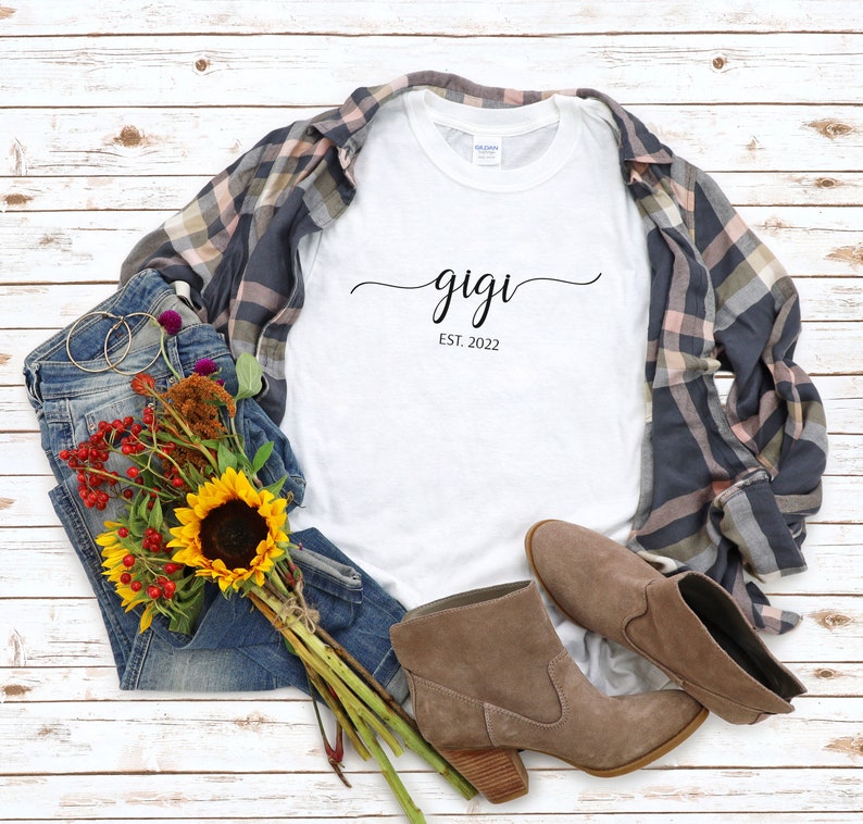 May include: White t-shirt with the text "gigi est. 2022" in black script font. The shirt is laid out on a white wood background with a pair of blue jeans, a plaid shirt, a bouquet of sunflowers and other flowers, and a pair of brown ankle boots.