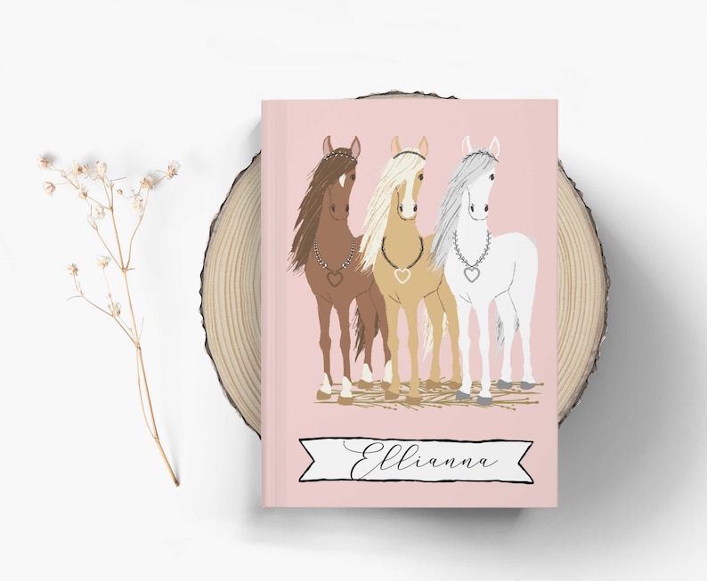 May include: A pink notebook with a white ribbon and the name "Ellianna" printed on it. The cover features a colorful illustration of three horses with hearts around their necks.