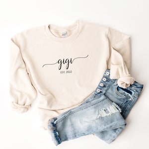 May include: A beige sweatshirt with the text "gigi est. 2022" in black script. The sweatshirt is paired with blue jeans.
