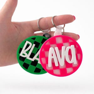 May include: Two round keychains with white text. One is green with a black and white checkered pattern and the letters "BLA". The other is pink with a white and pink checkered pattern and the letters "AVA".