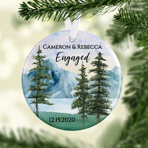 May include: Round white ceramic ornament with a watercolor painting of a mountain landscape with three pine trees. The text "Cameron & Rebecca Engaged 12.15.2020" is printed on the ornament.