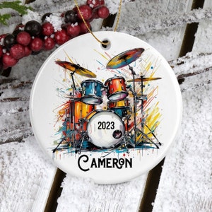 May include: A white ceramic Christmas ornament with a colorful illustration of a drum set. The drum set is surrounded by colorful paint splatters. The year 2023 is written on the bass drum. The name "CAMERON" is written below the drum set.