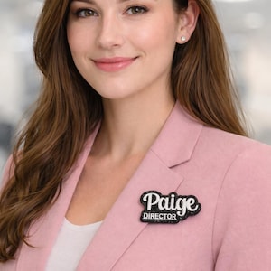 May include: A woman wearing a light pink blazer with a black and white name tag that says "Paige DIRECTOR". The name tag is a rectangular shape with a black glittery border and white text.