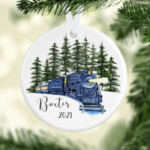 May include: A white ceramic Christmas ornament featuring a blue train with the words "Polar Express" on the side, surrounded by green pine trees. The ornament is personalized with the name "Baxter" and the year "2021".