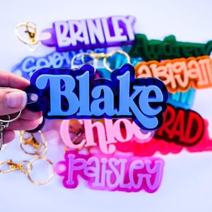 May include: A collection of colorful acrylic name keychains. The keychains feature names in various fonts and colors, including blue, pink, purple, green, and orange. Each keychain has a metal keyring attached. The name "Blake" is prominently displayed in blue.