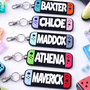 May include: Keychains with names: BAXTER, CHLOE, MADDOX, ATHENA, and MAVERICK. Each keychain is black with a colored border and white text. The keychains are designed to resemble a game controller. Silver key rings are attached.