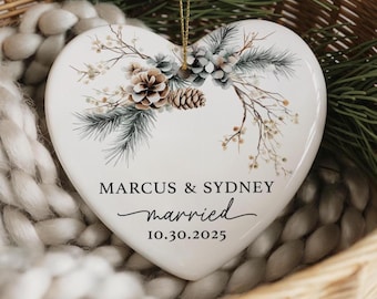 May include: White heart-shaped ceramic ornament with a botanical design of pinecones and foliage. The names "Marcus & Sydney", the word "married", and the date "10.30.2025" are printed on the ornament. A gold string is attached for hanging.