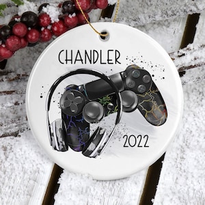 May include: White ceramic Christmas ornament with a black and white illustration of a video game controller and headphones. The ornament has the name "Chandler" and the year "2022" printed on it.