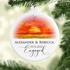 May include: A white ceramic ornament with a watercolor sunset over the ocean. The ornament is personalized with the names "Alexander & Rebecca" and the date "10.15.2021" with the word "Engaged" in a cursive font.