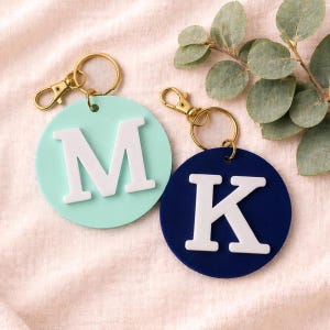 May include: Two round keychains with gold-colored hardware. One is light blue with a white "M", the other is navy blue with a white "K". The keychains are on a light pink surface with a sprig of greenery.