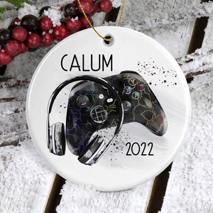 May include: White ceramic Christmas ornament with a black and white illustration of a video game controller and headphones. The ornament has the name "CALUM" and the year "2022" printed on it.