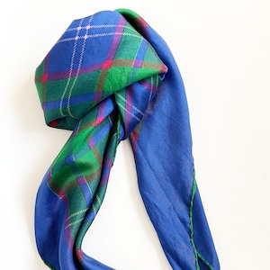 Puede incluir: Una bufanda de seda azul con un patrón de cuadros verdes, rosas y rojos. La bufanda está doblada y anudada.